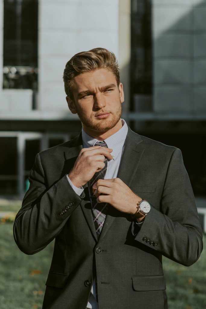 Serious confident male entrepreneur wearing classy suit and wristwatch holding tie while thoughtfully looking away
